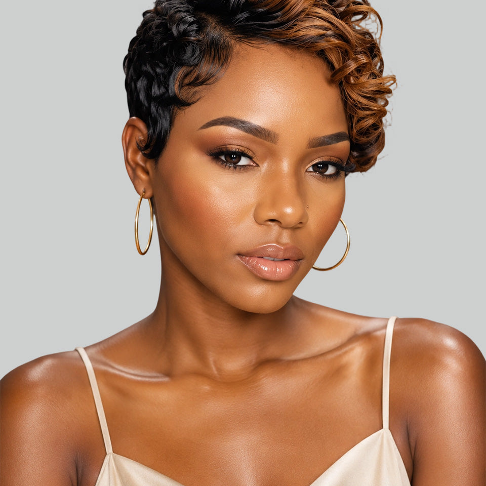 A woman with a short, curly, asymmetrical micro-lobed wig with dark brown and warm caramel highlights, wearing gold hoop earrings and a beige camisole against a solid gray background.