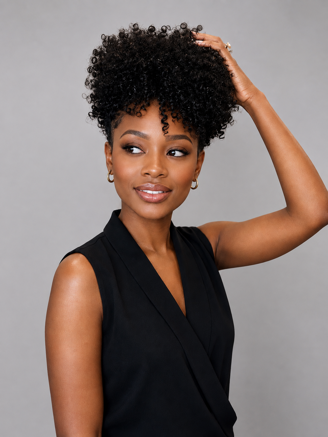 A woman with a voluminous, short, curly black wig or hairpiece posing against a solid gray background. She is wearing a black sleeveless top and small gold hoop earrings, with one hand touching the hair on top of her head.