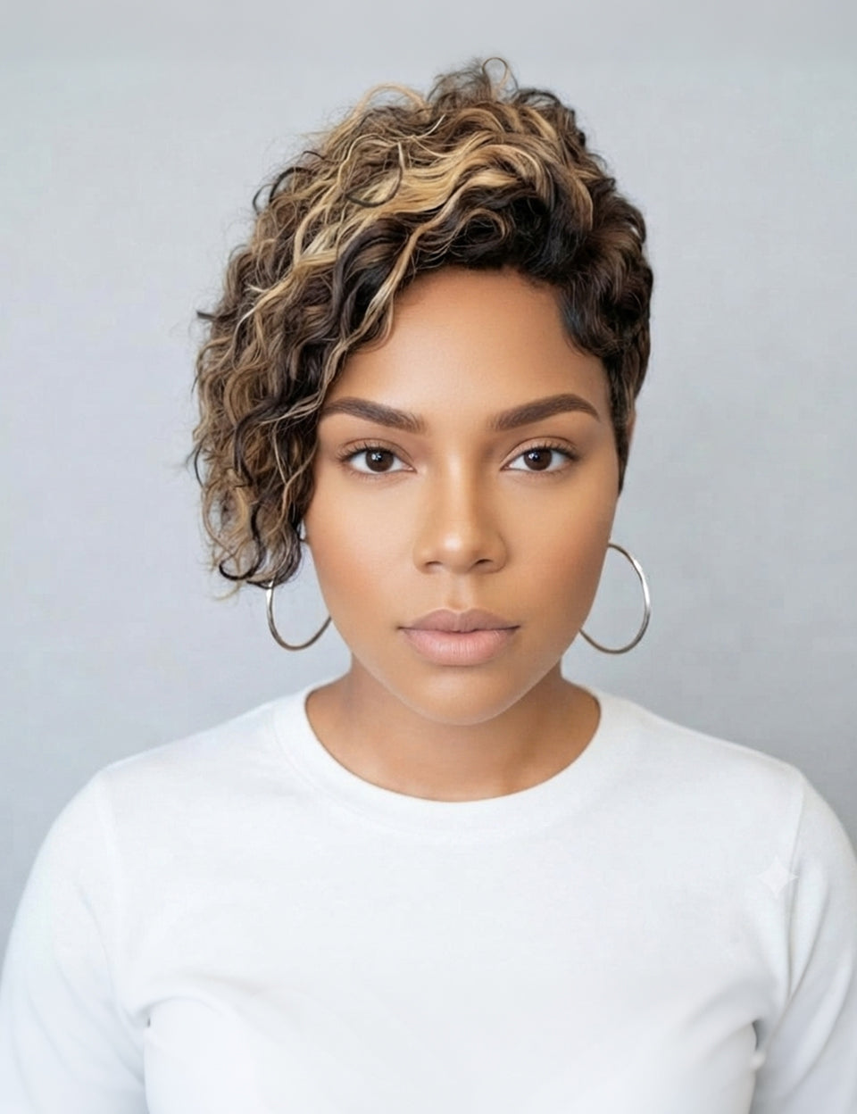 A woman with a short, curly wig featuring dark brown and blonde highlights, styled with large silver hoop earrings and a white long-sleeve top against a neutral gray background.