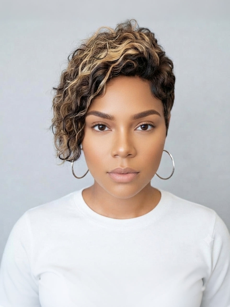 A woman with a short, curly wig featuring dark brown and blonde highlights, styled with large silver hoop earrings and a white long-sleeve top against a neutral gray background.