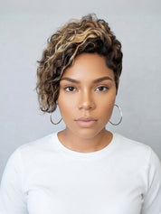 A woman with a short, curly wig featuring dark brown and blonde highlights, styled with large silver hoop earrings and a white long-sleeve top against a neutral gray background.