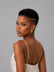 A woman with a short, textured black crop hairstyle is shown from the back over her shoulder, wearing a cream-colored spaghetti-strap top and small gold hoop earrings against a solid gray background.