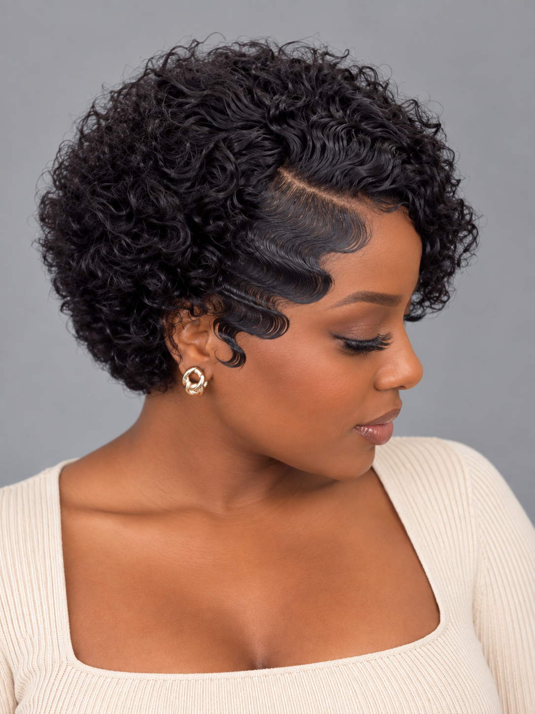 A woman with a short, curly black wig shown in profile against a plain gray studio background. The wig has defined waves and a clear side part, and she is wearing a cream ribbed top and a small gold earring.