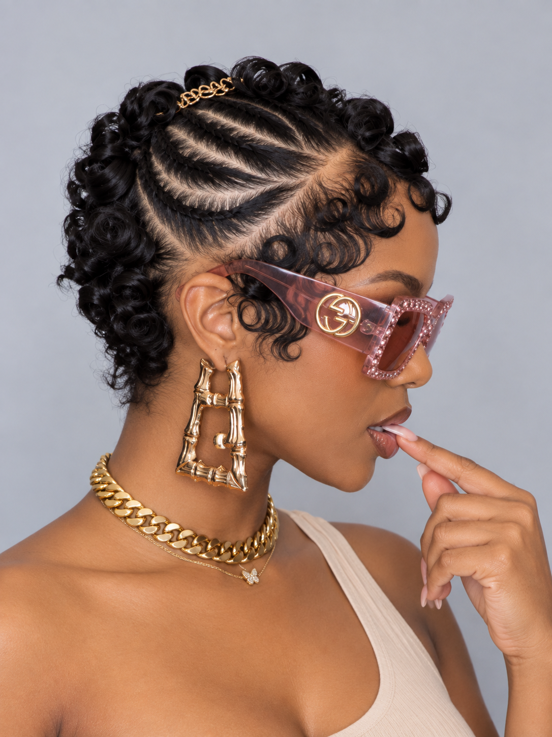 A woman in profile wearing Gucci sunglasses with a pink rhinestone frame and gold double-G logo, styled with a gold chain necklace and geometric earrings against a gray background.