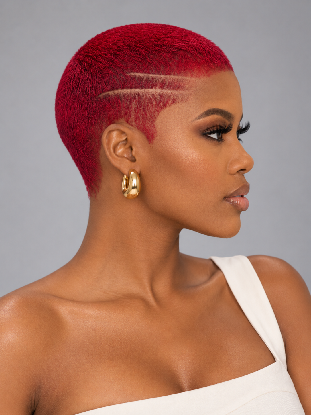 A close-up side profile of a woman with a vibrant red, micro-textured pixie cut and geometric side designs, wearing gold hoop earrings and a white strap top against a solid gray background.