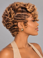 A woman shown in side profile wearing a short, curly wig with layered curls and a side-swept bang. The wig has a warm caramel brown base with golden blonde highlights, styled with a beige halter top against a plain gray studio background.
