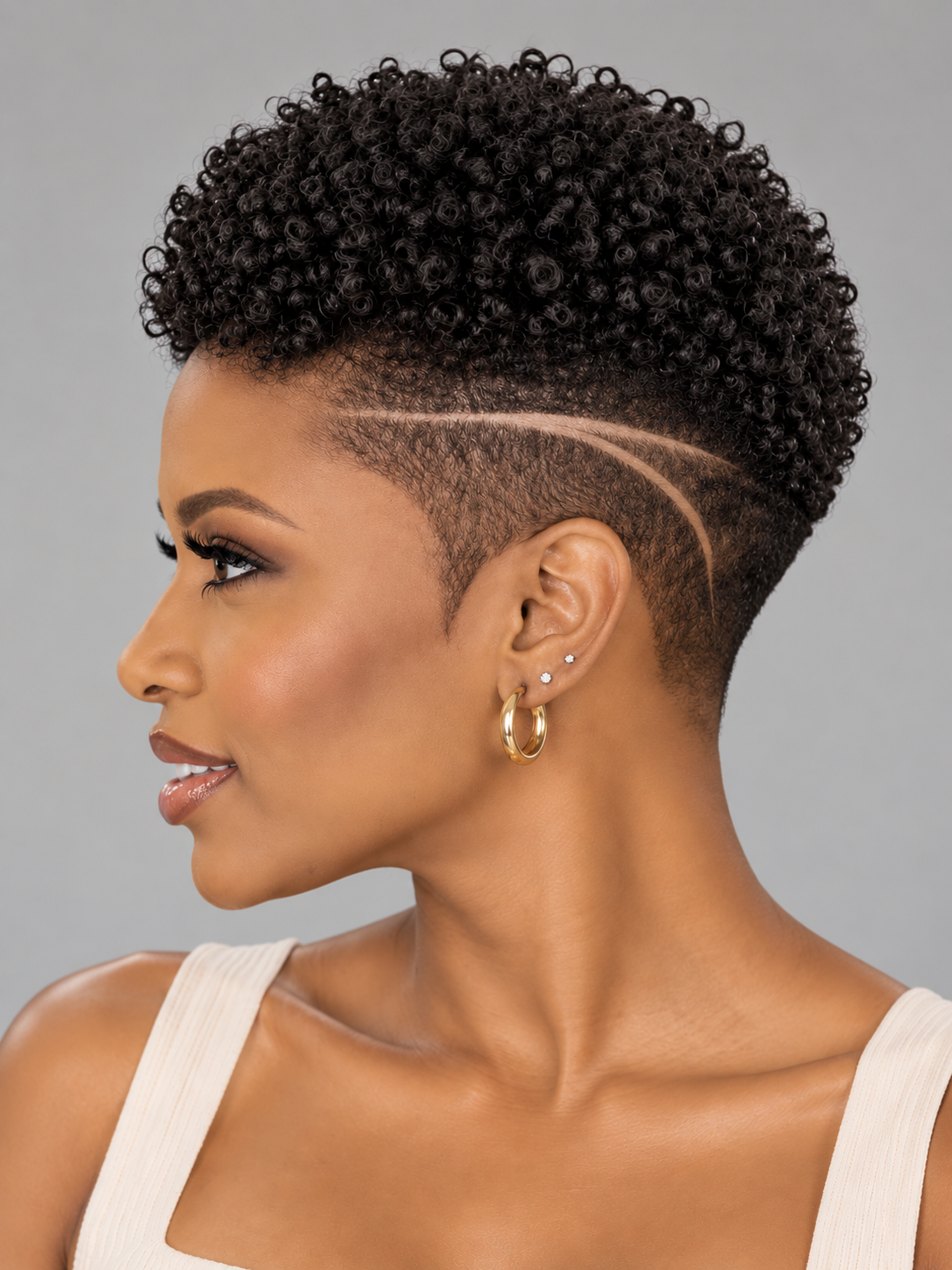 A woman shown in profile with a short, curly black haircut featuring a clean side razor shave and a curved line detail. She is wearing small gold hoop earrings against a plain light gray studio background.