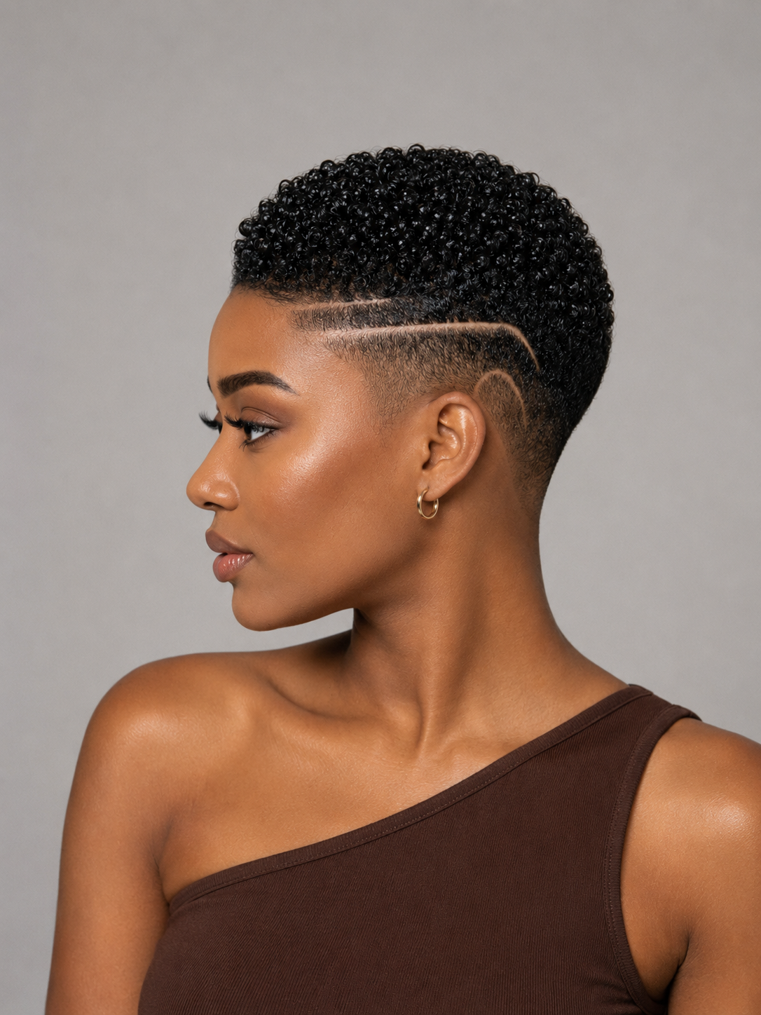 A woman shown in profile with a short, tightly styled black curly haircut and a clean side fade. She is wearing a dark brown one-shoulder top against a plain light gray studio background.