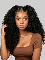 A woman with long, voluminous black curly hair styled with a braided hairline, wearing a brown scoop-neck top and a gold necklace, set against a solid gray background.