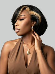 A woman with a short, voluminous black wig featuring highlighted blonde streaks along the front and sides, styled with a side part. She is wearing a brown halter top, gold hoop earrings, and a gold pendant necklace against a solid gray background.