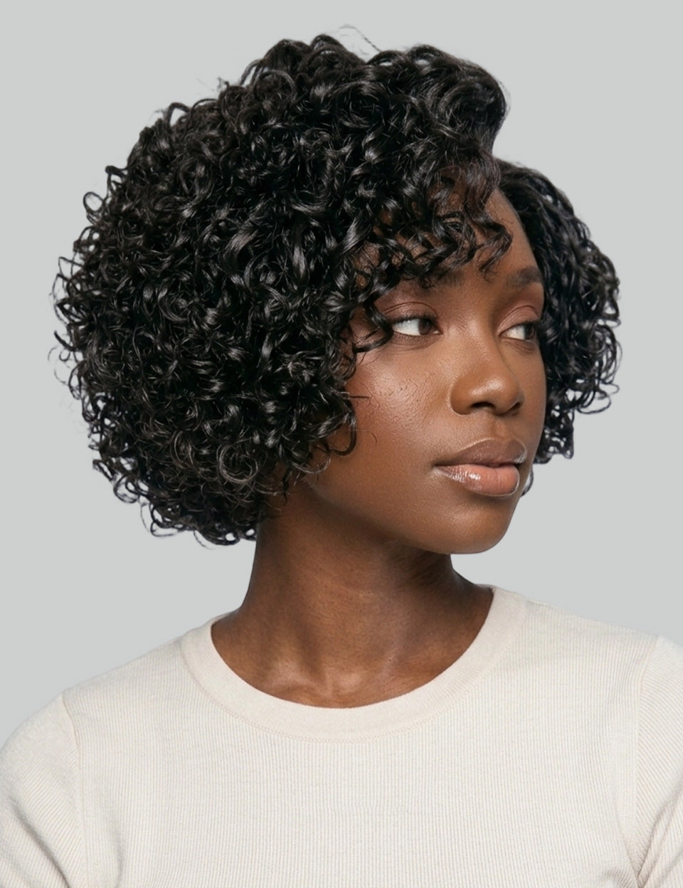 A woman wearing a short, curly black wig with a side-swept bang, shown from the chest up against a light gray studio background.