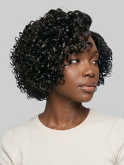 A woman wearing a short, curly black wig with a side-swept bang, shown from the chest up against a light gray studio background.