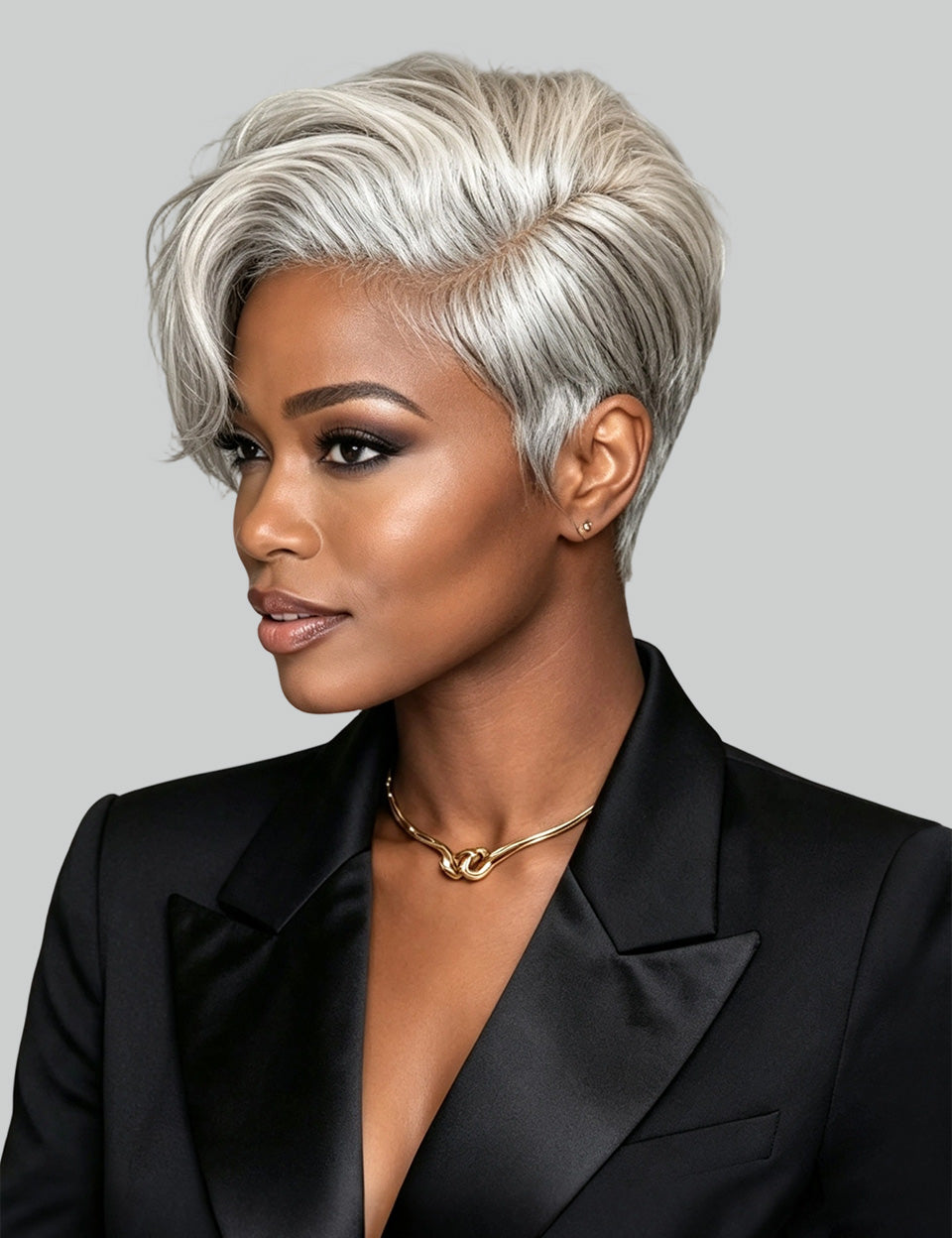 A woman with a short, layered silver-gray pixie cut hairstyle, wearing a black satin blazer and a gold chain necklace, shown in a three-quarter profile against a solid gray background.