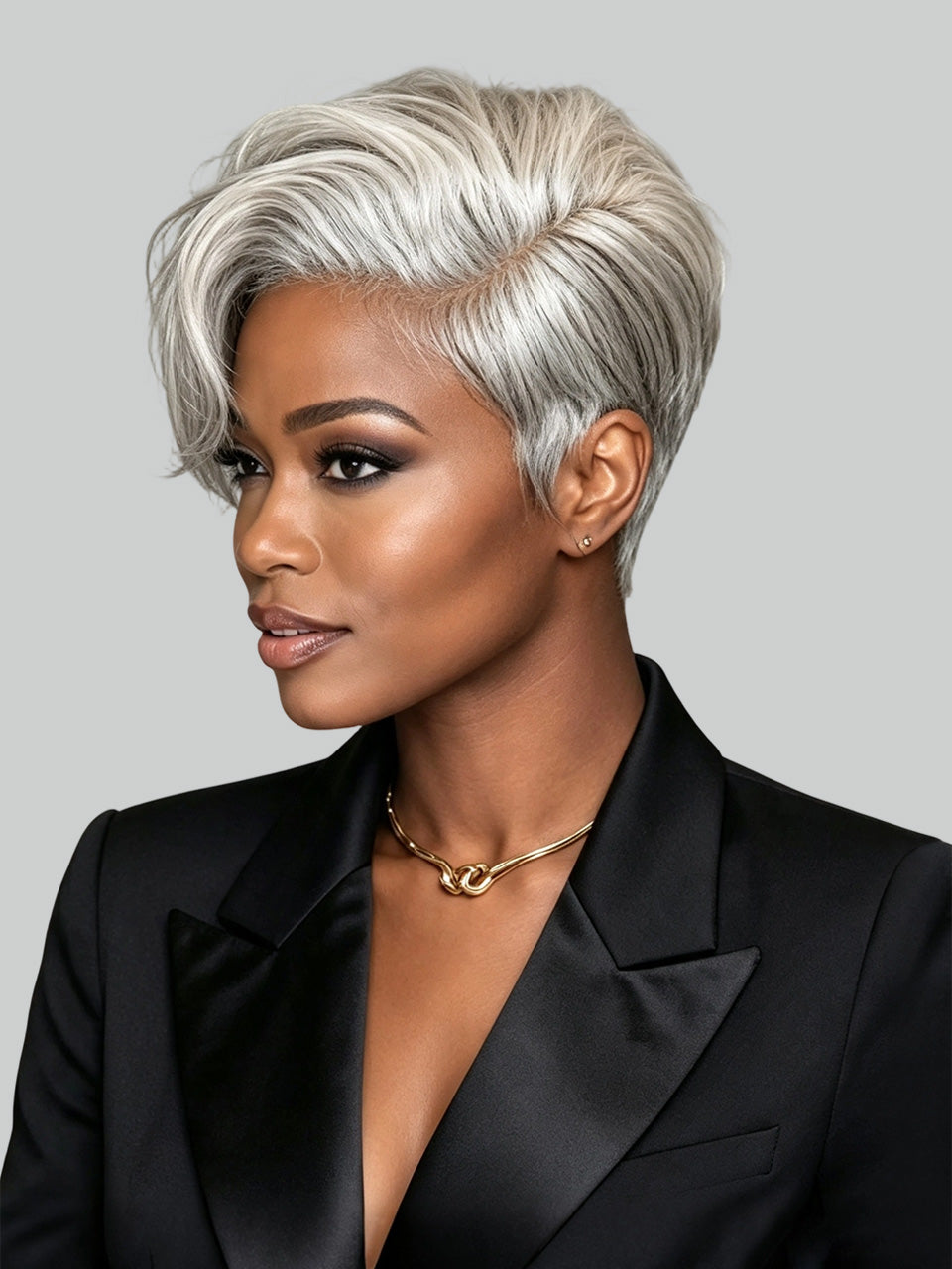 A woman with a short, layered silver-gray pixie cut hairstyle, wearing a black satin blazer and a gold chain necklace, shown in a three-quarter profile against a solid gray background.