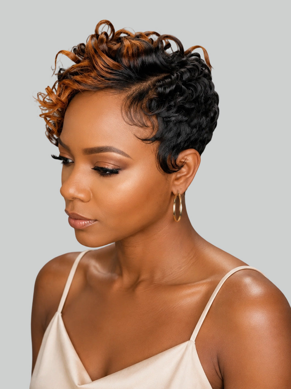 A woman with a short, curly wig featuring dark brown and warm caramel highlights, styled in a pixie cut. She is wearing a light beige spaghetti-strap top and gold hoop earrings against a solid light gray background.