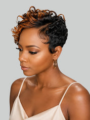 A woman with a short, curly wig featuring dark brown and warm caramel highlights, styled in a pixie cut. She is wearing a light beige spaghetti-strap top and gold hoop earrings against a solid light gray background.