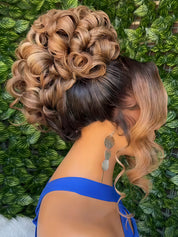 A woman shown from the back wearing an elaborate updo hairstyle with layered blonde and dark brown tones, a bright blue strap visible at the shoulder, and long dangling earrings, set against a green leafy background.