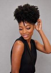 A woman with a voluminous, tightly curled black afro hairstyle wearing a black sleeveless top, posing against a solid gray background.