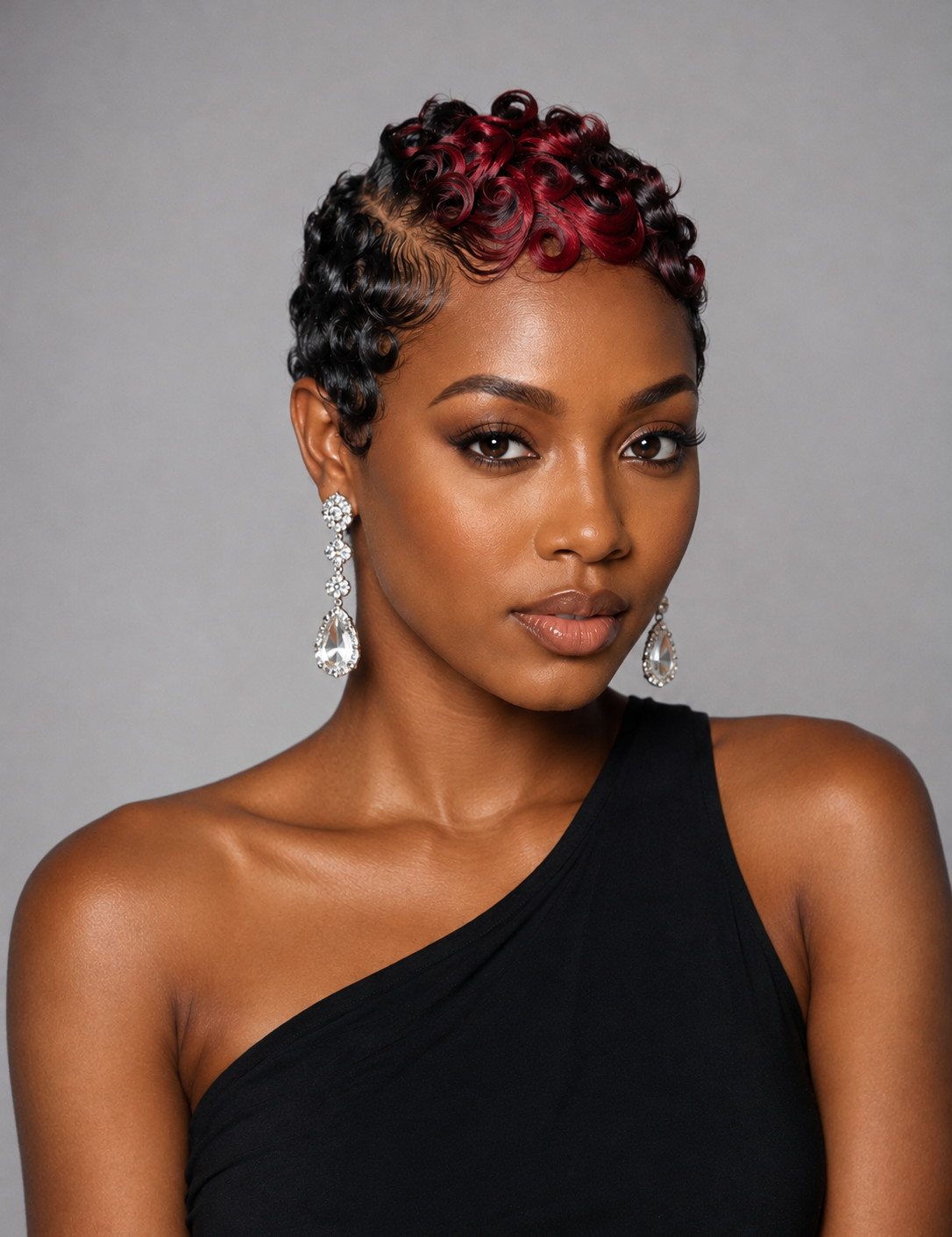 A woman with a short, curled hairstyle featuring dark brown roots and deep red highlights, wearing a black one-shoulder top and dangling earrings, set against a solid gray background.