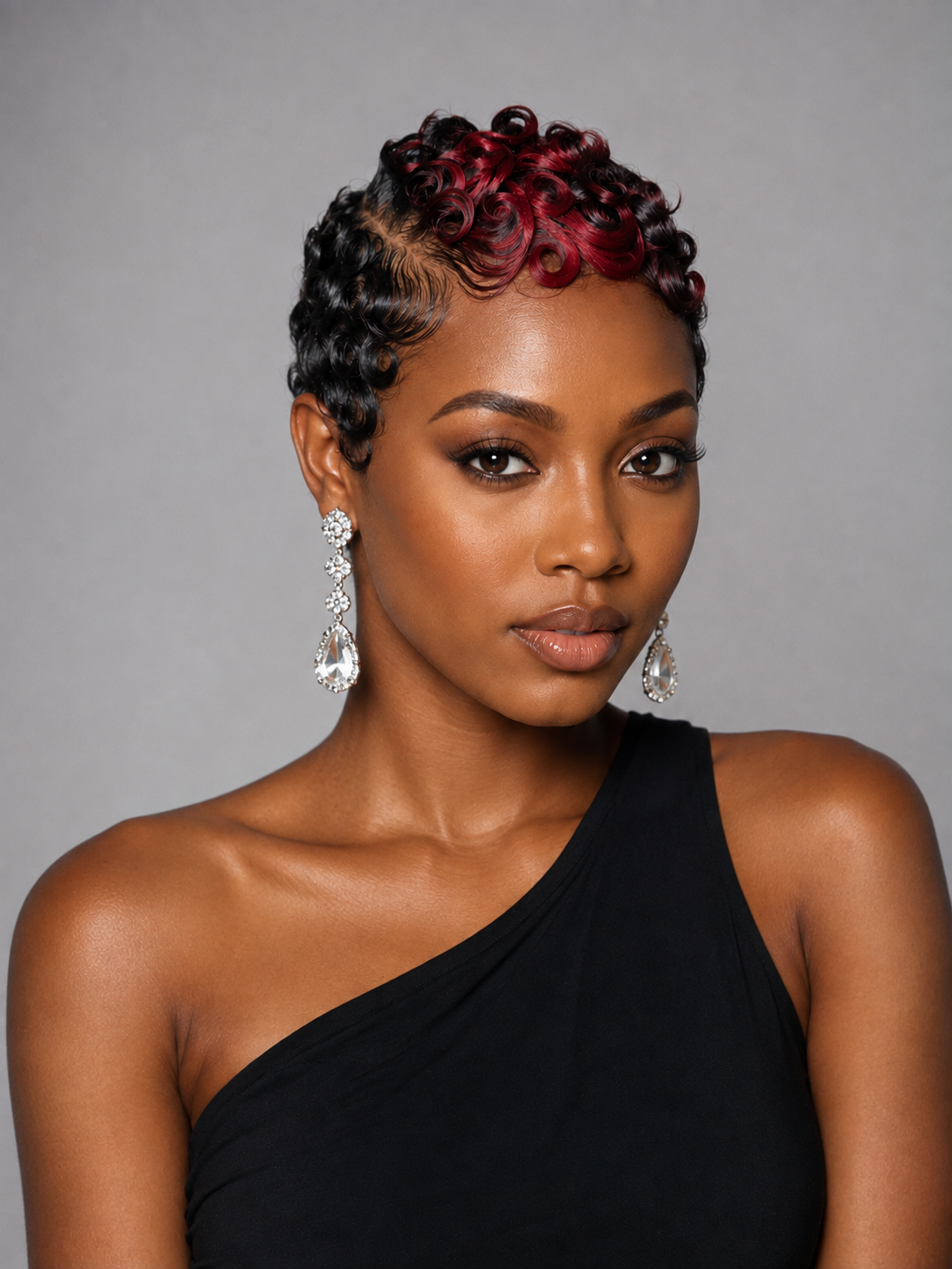 A woman with a short, curled hairstyle featuring dark brown roots and deep red highlights, wearing a black one-shoulder top and dangling earrings, set against a solid gray background.