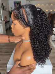 A mannequin bust wearing a long, dark curly wig with a side part and braided fringe, shown indoors with a person's hands adjusting the hair. The wig is the product being demonstrated on a mannequin, so this is best classified as Product/Packaging rather than Lifestyle because mannequins do not count as people under the schema.