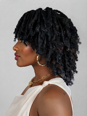 A woman shown in profile wearing a voluminous, textured black dreadlock hairstyle. She is wearing a white draped top, gold hoop earrings, and a gold chain necklace against a plain light gray studio background.