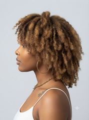 A woman shown in profile with voluminous, tightly curled natural hair. She is wearing a white spaghetti-strap top, large gold hoop earrings, and a delicate gold chain necklace against a plain light gray studio background.