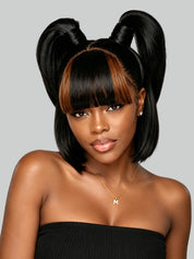 A woman with a black bob haircut, straight-cut bangs, and two high pigtails styled with warm brown highlights, wearing a black strapless top against a light gray background.