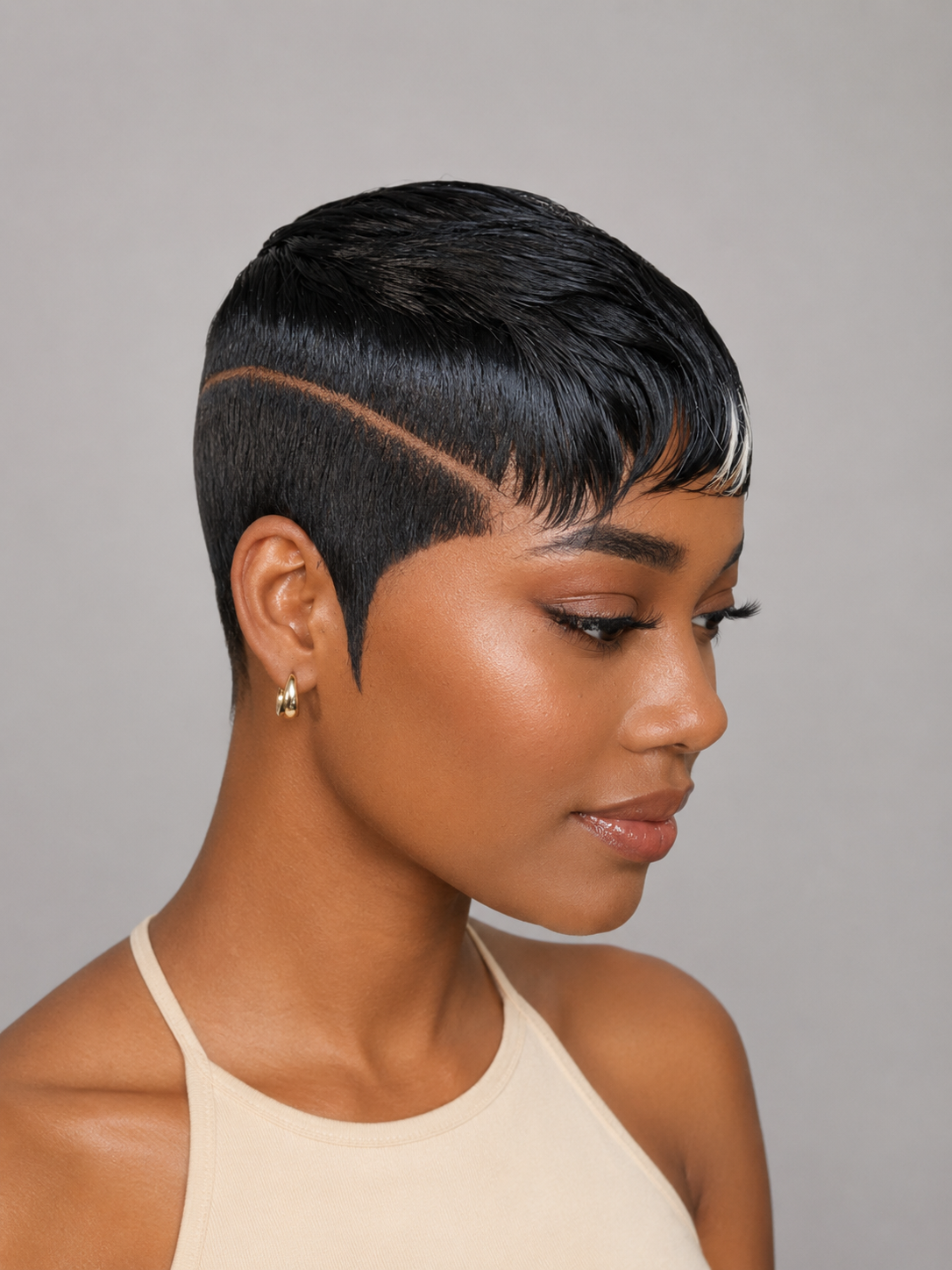 A woman shown in profile wearing a short, dark wig with a sharp side undercut and a fine part line. The wig is styled with a sleek, glossy finish against a plain gray studio background.