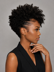 A woman with a short, tightly curled black afro hairstyle is shown in profile against a plain gray studio background. She is wearing a black sleeveless top and small gold hoop earrings, with her hand resting near her chin.