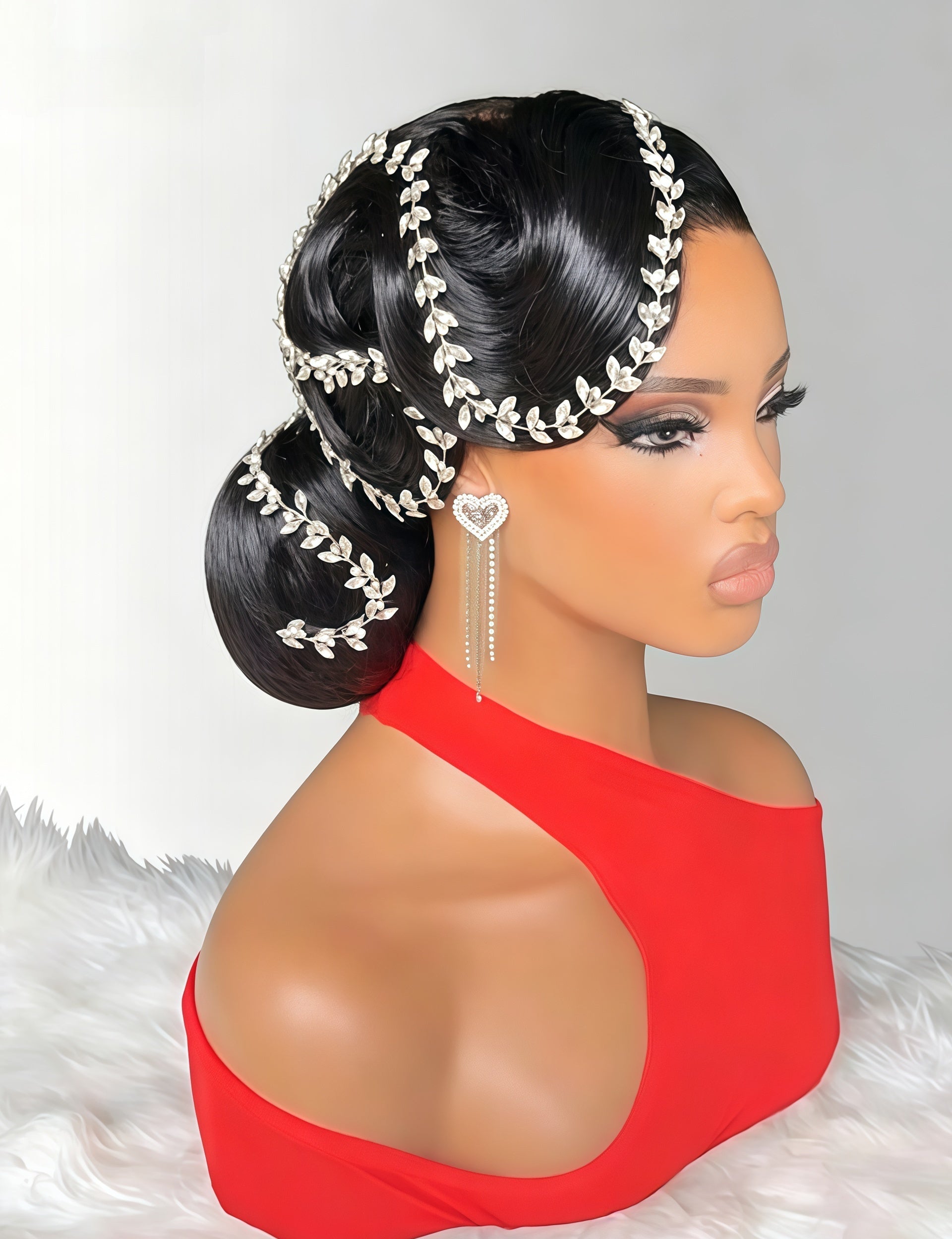 A mannequin bust with dark hair styled in an elegant updo, decorated with a silver leaf-patterned headpiece and matching dangling earrings, wearing a bright red one-shoulder top against a light gray background with a white faux-fur surface.