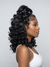 A woman in profile wearing a black curly half-updo wig with a small silver hair accessory, shown against a light gray studio background.