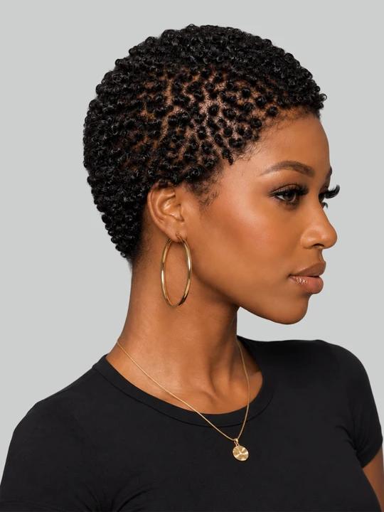 A woman in profile wearing a short, tightly curled black hairstyle, large gold hoop earrings, and a delicate gold necklace against a solid gray background.