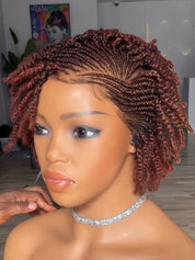 A mannequin head wearing a short, styled twist-out wig in a warm reddish-brown shade, shown indoors with a hand adjusting the hair. The background includes a framed poster and light-colored walls.