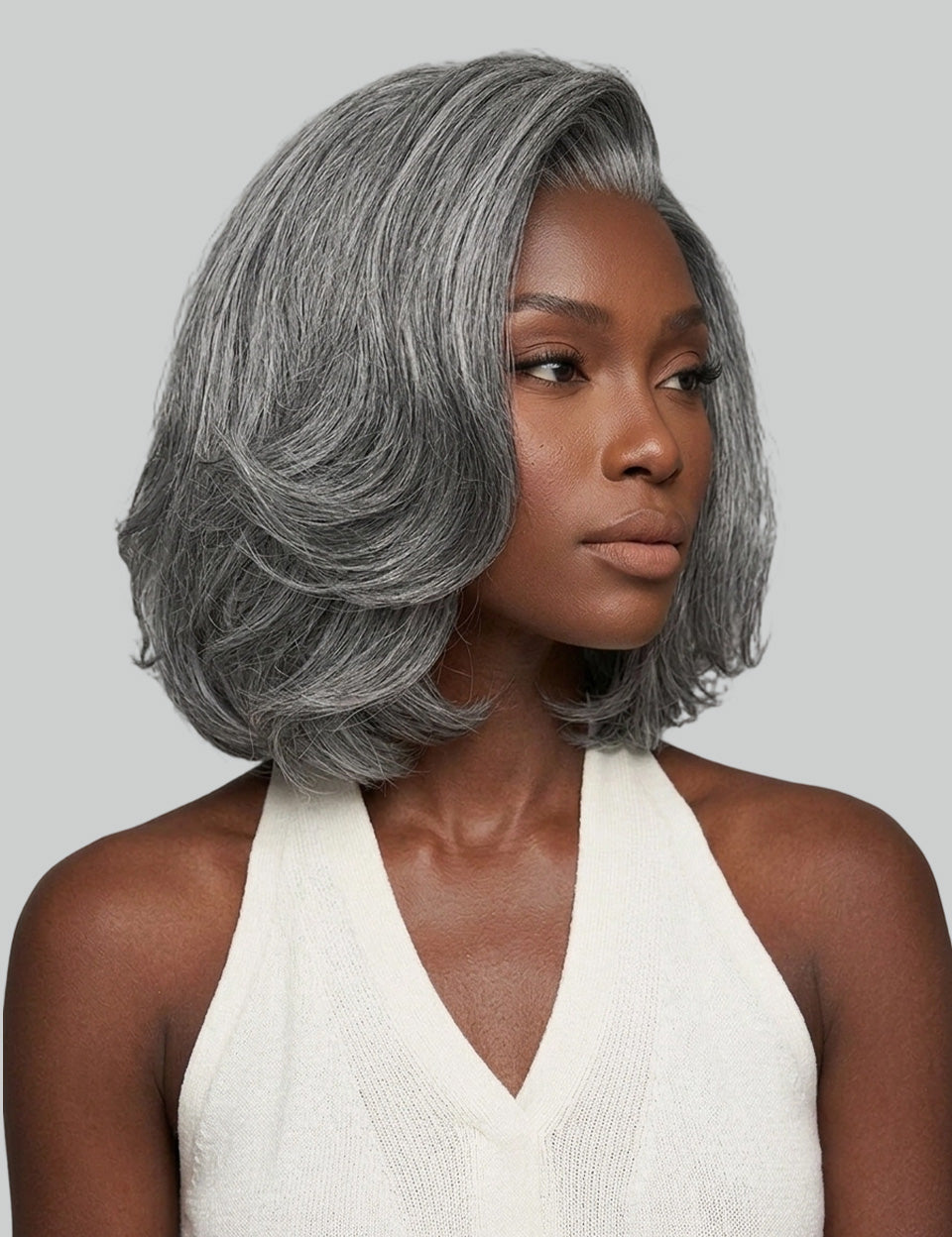 A woman wearing a short, layered gray wig with subtle silver highlights, styled in a bob length with soft waves. She is wearing a white halter top against a solid light gray background.