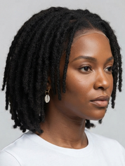 A close-up portrait of a woman with short, textured black dreadlocks, wearing a white crew-neck t-shirt and a small silver shell earring, set against a plain light gray background.
