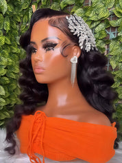 A mannequin bust wearing a long, dark, wavy wig with side parting, styled with a silver rhinestone hair accessory and dangling earrings, against a green leafy background.