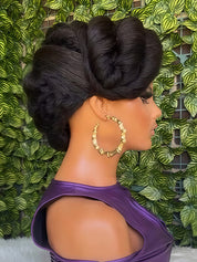 A side-profile view of a mannequin or model with an elaborate dark brown updo hairstyle, wearing large gold hoop earrings and a purple top, set against a green leaf-patterned background.