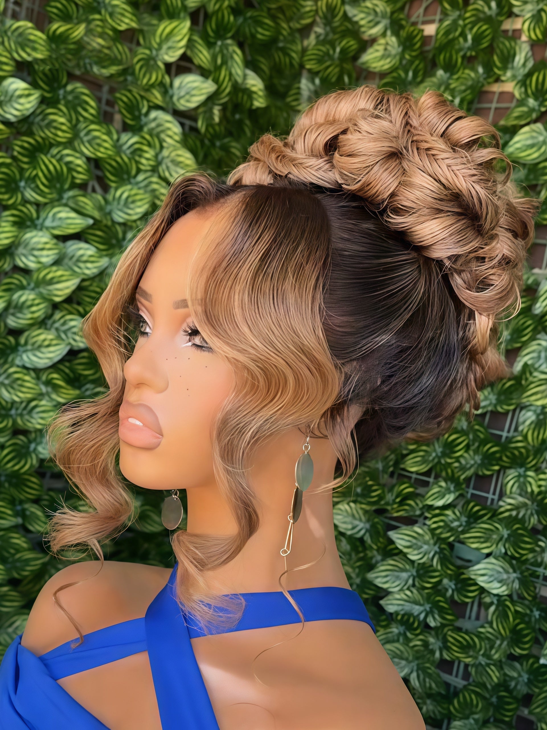 A mannequin or model head wearing a styled wig with wavy medium brown and blonde ombre sections, featuring a high braided updo and side-swept bangs, set against a green leaf wall background.