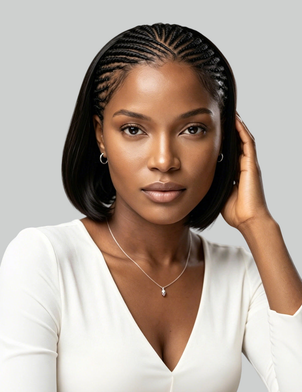 A woman with a short black bob hairstyle and cornrow braids, wearing a white V-neck top, a delicate silver necklace, and small hoop earrings, posing against a solid gray background.