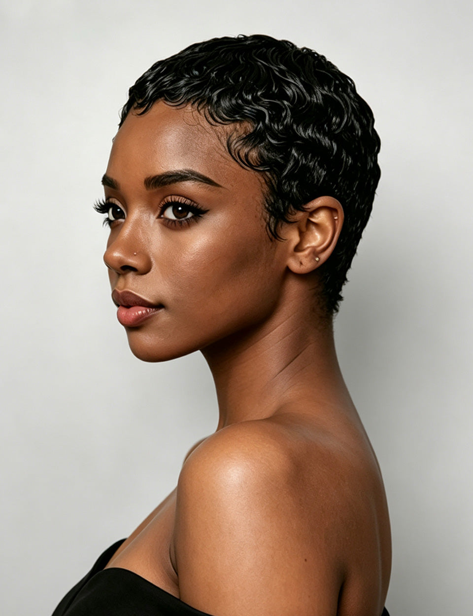 A woman wearing a short, voluminous curly wig in black, styled with a high puff on top, paired with a white V-neck t-shirt against a solid gray studio background.