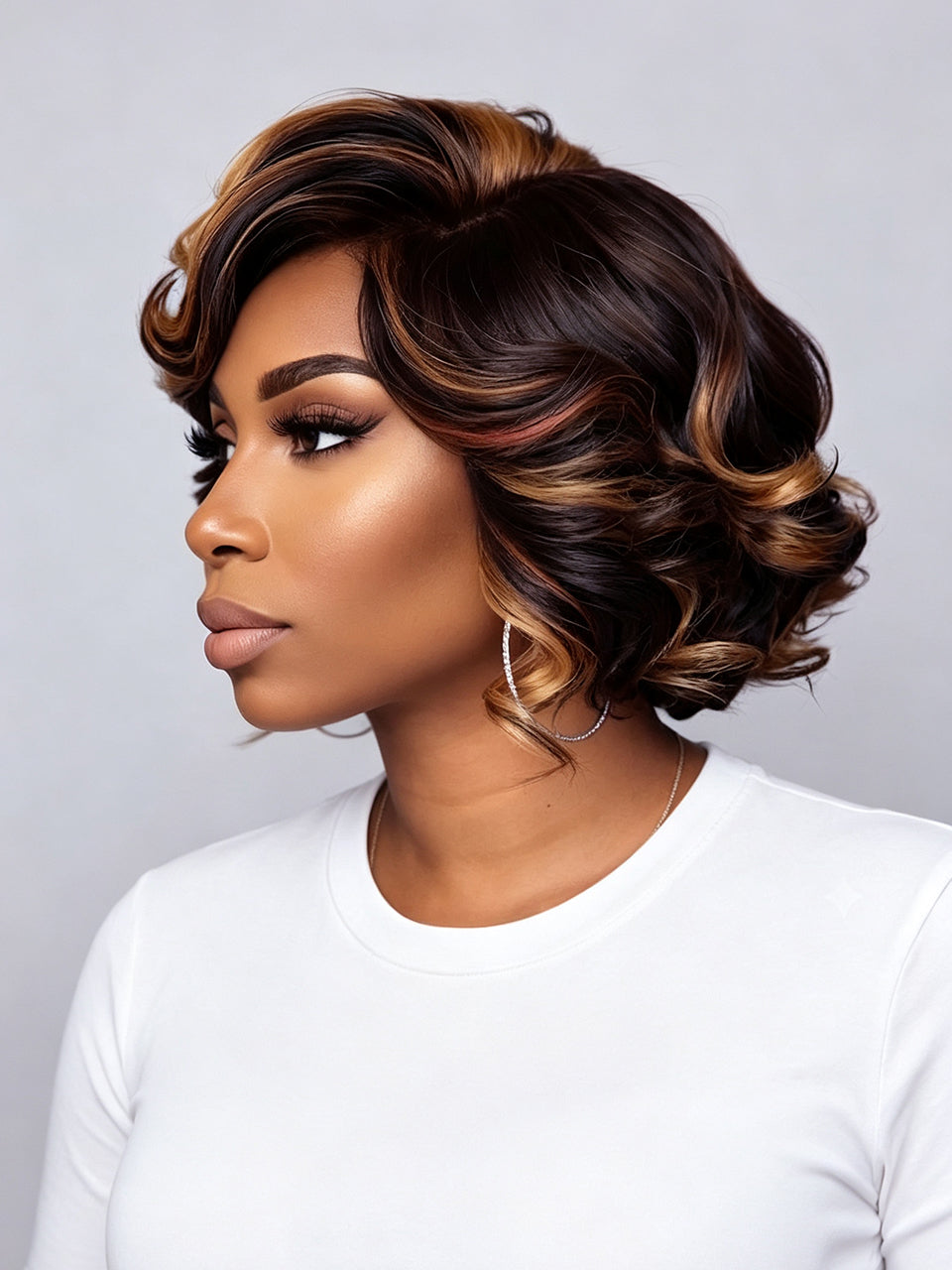 A woman shown in profile wearing a short, styled wig with deep brown and warm caramel-blonde highlights, paired with a plain white top against a light gray studio background.