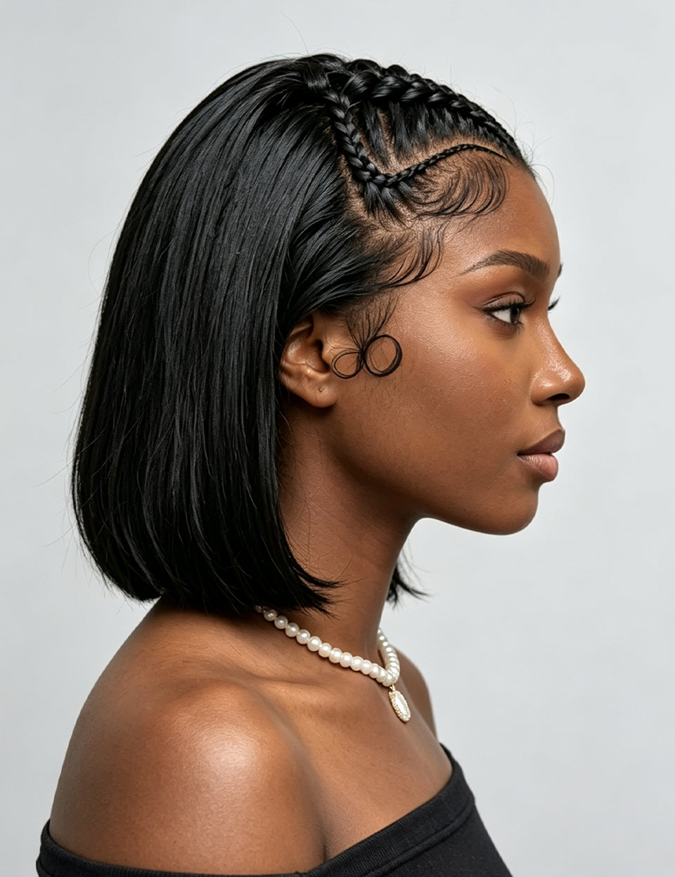 A woman in profile wearing a short, straight black wig with braided detailing along the crown and a pearl necklace against a light gray background.
