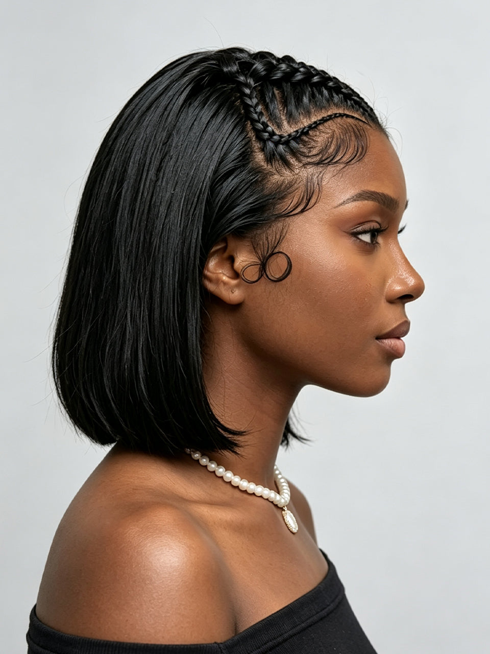 A woman in profile wearing a short, straight black wig with braided detailing along the crown and a pearl necklace against a light gray background.