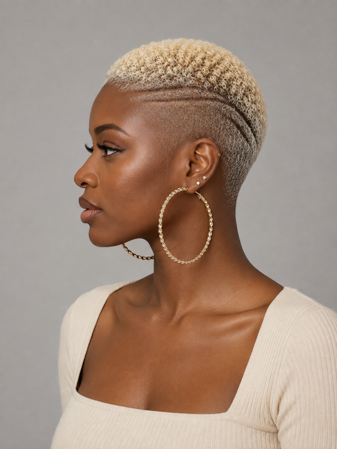 A woman in profile wearing large gold hoop earrings with a beaded texture, a short textured blonde haircut, and a cream ribbed top against a solid gray background.