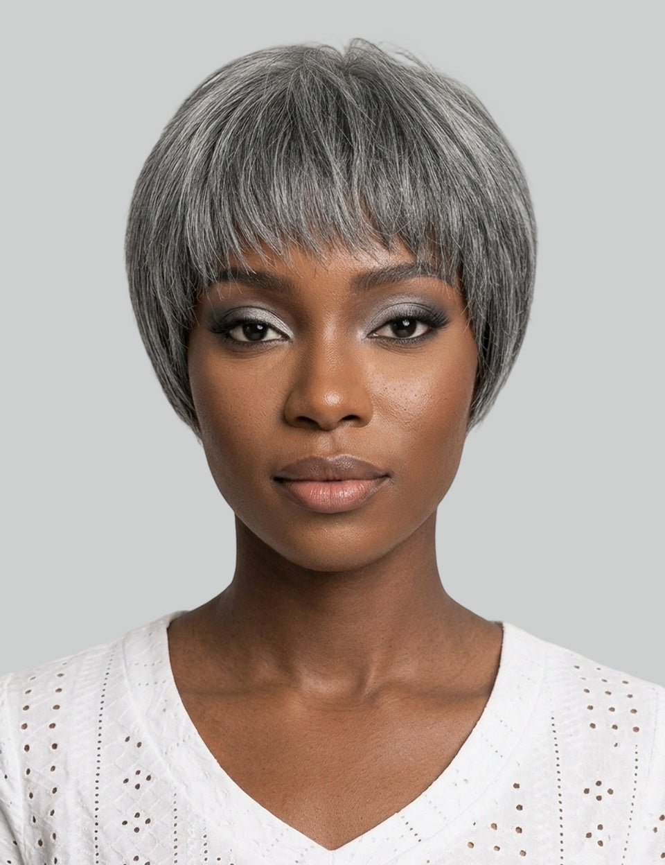 A woman with a short, layered gray wig and silver-gray eyeshadow, wearing a white eyelet V-neck top against a solid gray background.