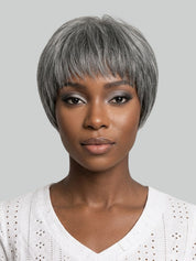A woman with a short, layered gray wig and silver-gray eyeshadow, wearing a white eyelet V-neck top against a solid gray background.