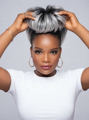 A woman with a voluminous, high-top updo hairstyle featuring a silver and black ombre pattern, wearing a white t-shirt and gold hoop earrings against a light gray background.