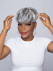 A woman wearing a short, layered silver and gray wig with bangs, posing against a light gray background while adjusting her hair with both hands.