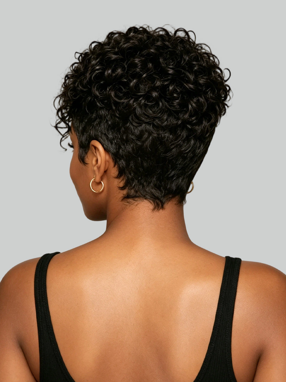 Rear view of a woman with short, curly black hair, wearing a black spaghetti-strap top and a gold hoop earring against a solid gray background.